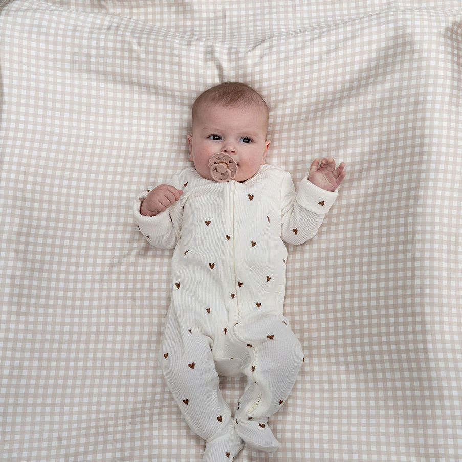 A baby lying on a cream and white gingham blanket on a light wood floor, wearing a white footed sleeper with small brown heart prints.