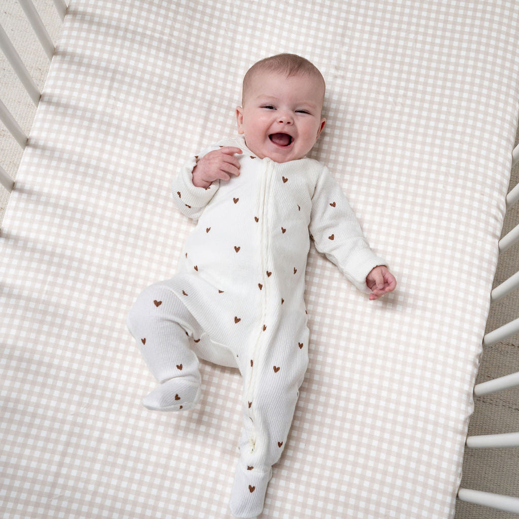A smiling baby lies on their back inside a white crib, wearing a white footed sleeper with small brown heart prints. The crib is fitted with a beige and white gingham check sheet.